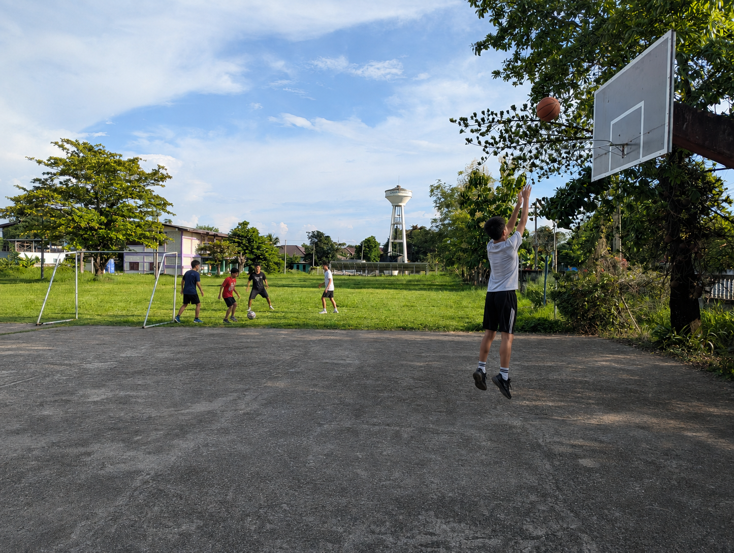 A learner practicing basketball while a small group plays football on the grass